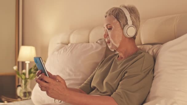 Mujer asiática mayor que usa mascarilla de hoja facial y auriculares inalámbricos navegando en el teléfono inteligente mientras se relaja en la cama tarde en la noche - Metraje, vídeo