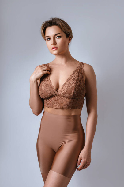 Portrait of young attractive woman wearing brown lace bra and high waist shaping briefs against gray background. - Foto, immagini