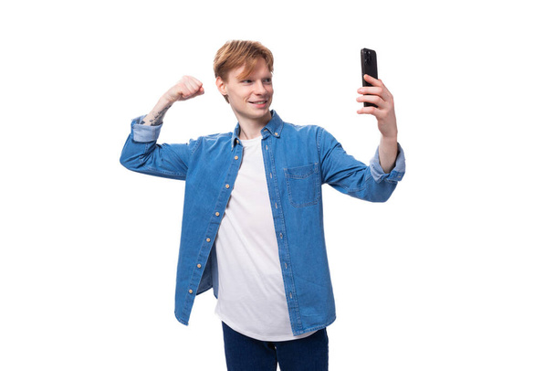 Ένας νεαρός κοκκινομάλλης με μπλε τζιν πουκάμισο βγάζει μια σέλφι στο τηλέφωνο στο βάθος με φωτοτυπίες.. - Φωτογραφία, εικόνα