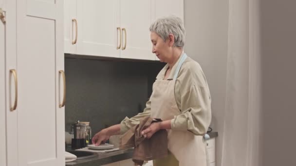Tiro medio de mujer asiática mayor de pelo gris secando platos con toalla y poniéndolos en el estante más bajo del gabinete de cocina blanco - Metraje, vídeo