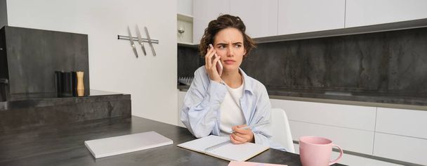 Portrait of woman working from home, calling someone on mobile phone, looking confused while holding pen and trying to write down something, sitting with puzzled face in kitchen. - Photo, Image