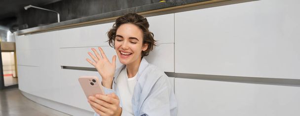 Χαριτωμένη μελαχρινή χαμογελά στην κάμερα smartphone, κύματα χέρι, λέει γεια, συνδέεται με συνομιλίες βίντεο, μιλάει σε κάποιον, κάθεται στο πάτωμα στην κουζίνα. - Φωτογραφία, εικόνα