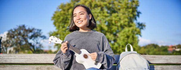 Preciosa joven sentada con mochila en el banco en el soleado parque, toca la guitarra ukelele y canta. Estilo de vida - Foto, Imagen