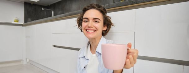 Positivo, sorridente giovane donna siede sul pavimento della cucina, si estende mano con la tazza, ti offre tazza con caffè o tè, gode la sua bevanda. - Foto, immagini