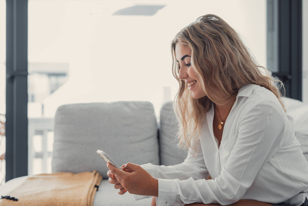 Портрет молодой привлекательной блондинки, пользующейся телефоном на диване, расслабляющей серфинг в сети дома. Расслабляющий стиль жизни онлайн - Фото, изображение
