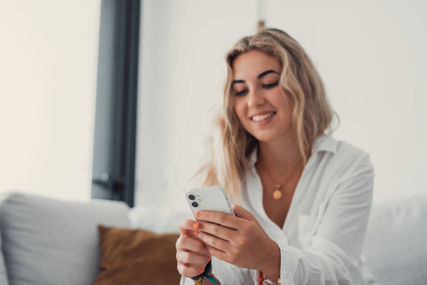 Портрет молодой привлекательной блондинки, пользующейся телефоном на диване, расслабляющей серфинг в сети дома. Расслабляющий стиль жизни онлайн - Фото, изображение