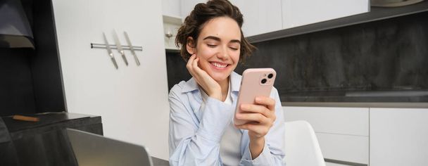 Imagen de una mujer joven usando su smartphone en casa. Chica se sienta con el teléfono móvil en la cocina y sonríe. - Foto, imagen