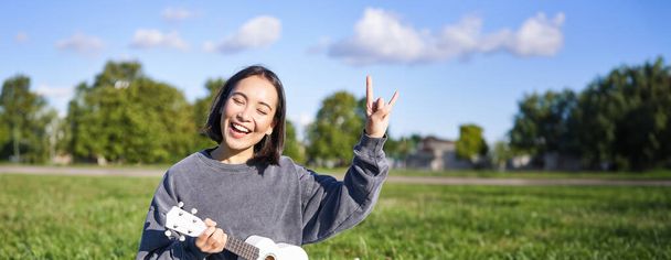 Szczęśliwa azjatycka dziewczyna gra ukulele w parku, pokazując rock n roll, heavy metal rogi znak i uśmiech, zabawy na świeżym powietrzu. - Zdjęcie, obraz