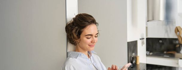Porträt einer lächelnden, schönen Frau, die aufs Smartphone schaut, zu Hause in der Nähe der Küche steht, Nachrichten checkt, mit dem Handy chattet. Kopierraum - Foto, Bild