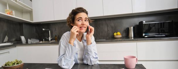 Portrait de femme pensant en parlant sur téléphone portable, assis avec smartphone dans la cuisine à la maison et crayon mordant avec visage confus. - Photo, image