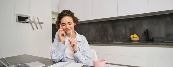 Bild einer Frau, die zu Hause arbeitet, telefoniert, mit dem Smartphone sitzt, von Papieren umgeben ist, Hausaufgaben macht und mit jemandem spricht. - Foto, Bild