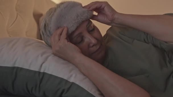 Klidný senior asijské žena ležící na polštáři pod měkkou přikrývku, na sobě hedvábí spací maska a usínání v útulné posteli v noci - Záběry, video