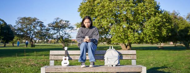 Kaunis brunette tyttö penkillä puistossa, istuu ukulele ja reppu, tilalla älypuhelin, mobiilisovelluksella. - Valokuva, kuva