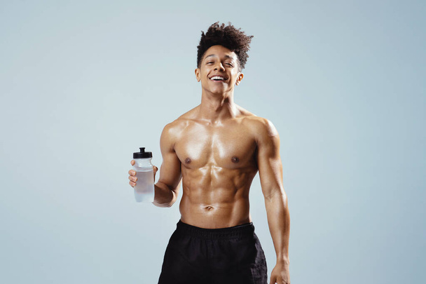 Workout Hydration. Cheerful Athletic Young Man Holds Bottle Of Water And Smiles To Camera On Gray Background. Studio Shot Of Handsome Muscular Fitness Guy With Shirtless Torso Hydrating After Sport - Fotografie, Obrázek