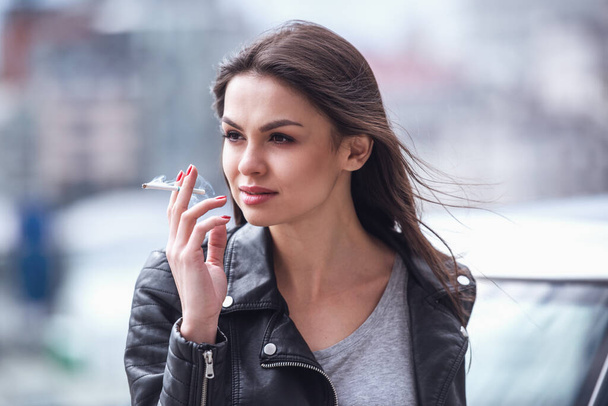 Hermosa chica en chaqueta de cuero está fumando y mirando hacia otro lado mientras se apoya en su coche
 - Foto, imagen