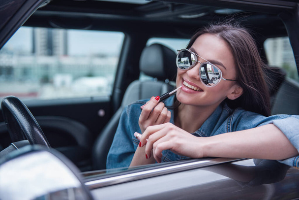 Beautiful girl in jean jacket and sun glasses is applying lip gloss and smiling while sitting in her car - Photo, Image