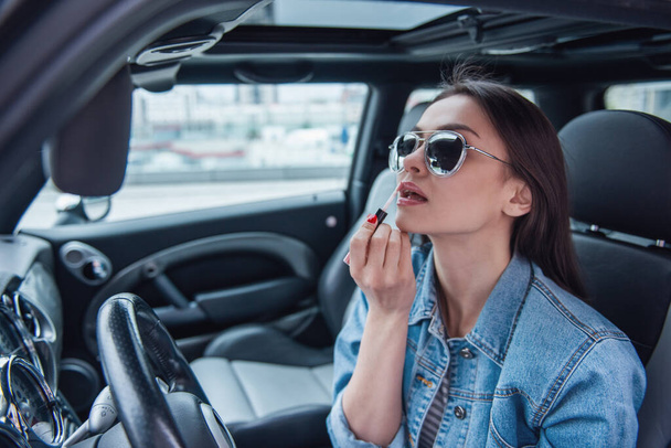 Beautiful girl in jean jacket and sun glasses is applying lip gloss while sitting in her car - Photo, Image