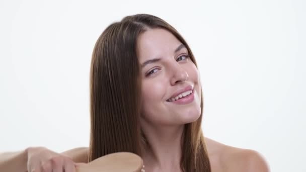 Woman attentively cares for hair, gently gliding a comb through locks with a focused expression against a pristine white backdrop. Her graceful movements reflect a sense of self-love and care. - Footage, Video