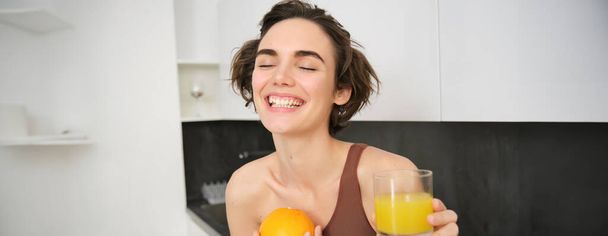 Healthy lifestyle and sport. Beautiful smiling woman, drinking fresh orange juice and holding fruit in her hand, enjoying her vitamin drink after workout at home, standing in kitchen. - Photo, Image