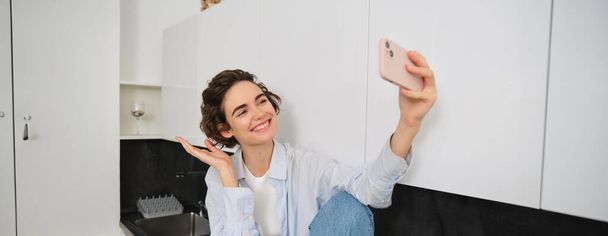 Beautiful woman takes selfie on smartphone, posing in kitchen and smiling at mobile camera for photo. - Photo, Image