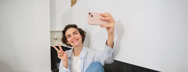 Portrait de belle fille brune, prend selfie sur smartphone dans sa cuisine, posant pour photo sur téléphone portable. - Photo, image