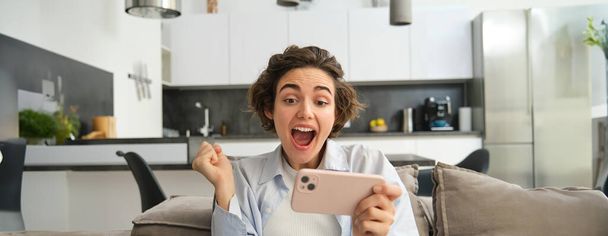 Retrato de mulher jogando jogos de celular, assistindo vídeo com rosto animado, usando smartphone para lazer, senta-se no sofá em casa. Estilo de vida e conceito de pessoas - Foto, Imagem