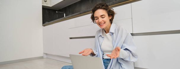 Portrait de jeune fille freelance se connecte à une réunion en ligne, chats vidéo sur ordinateur portable, se trouve sur le sol de la cuisine et aime sa conversation. - Photo, image