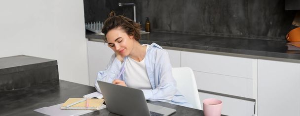 Wizerunek młodej produktywnej kobiety pracuje w domu, uczy się na odległość, uczy się online na laptopie w kuchni, zapisuje informacje, ogląda webinarium, uczestniczy w spotkaniu zdalnie. - Zdjęcie, obraz