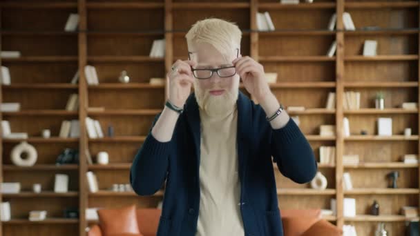 Portret van een albino man met een bril in de bibliotheek. Vermoeide zakenman die elkaars hand kruist in het kantoor. Slimme vent met doordachte gezichtsuitdrukking. Jonge ondernemer uitgeput na woking dag - Video
