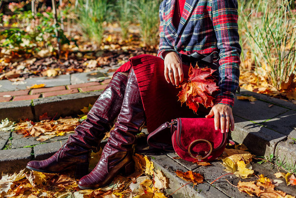 Primer plano de botas altas de piel roja borgoña. Mujer de moda con elegante falda de chaqueta a cuadros sentado en el parque de otoño con bolso entre las hojas caídas - Foto, Imagen