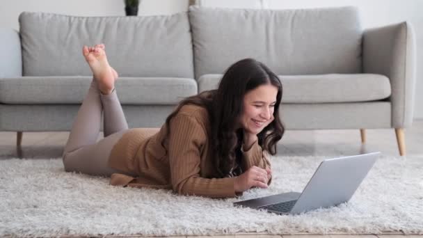Dans un moment de pure joie, une femme caucasienne positive est allongée sur le sol, partageant des rires chaleureux tout en utilisant un ordinateur portable. Son visage est illuminé d'un vrai bonheur, des rires résonnent dans la pièce. - Séquence, vidéo