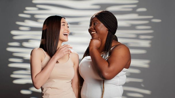 Curvy young adult telling secret to asian lady on camera, acting sensual and positive in studio. Diverse beauty models advertising body positivity and self confidence, having fun. - Photo, Image