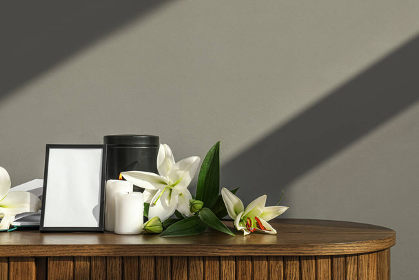 Marco fúnebre en blanco, urna mortuoria, velas encendidas y flores de lirio en la mesa de madera cerca de la pared gris - Foto, imagen
