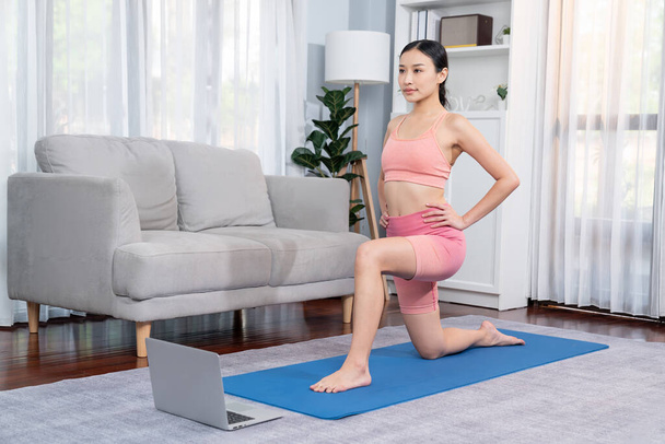 Mujer asiática en ropa deportiva haciendo ejercicio de yoga en la alfombra de fitness como su rutina de entrenamiento en casa. Cuidado del cuerpo saludable estilo de vida mujer viendo video de yoga en línea en el ordenador portátil. Vigoroso - Foto, Imagen