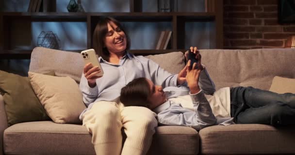 Sohn und Mutter verbringen einen ruhigen Moment auf dem Sofa, beide in Telefone vertieft. Das sanfte Leuchten der Bildschirme erhellt Gesichter und unterstreicht den Fokus und die gemeinsame Gelassenheit des Augenblicks. - Filmmaterial, Video