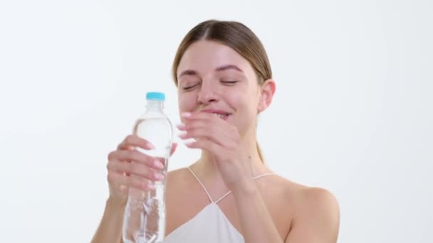 Vidám, kaukázusi származású nő mosolyog, miközben könnyedén kinyitja a vizes palackot, és frissítő kortyot iszik. A boldogság sugárzik a fehér háttérben, megragadja a tiszta öröm pillanatát.. - Felvétel, videó