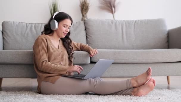 Žena pohodlně sedí na podlaze, nosí sluchátka a poslouchá hudbu a zároveň se věnuje laptopu. Její tvář odráží potěšení z melodií. - Záběry, video