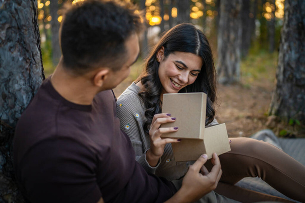 Two people young adult caucasian man and woman couple boyfriend and girlfriend or husband and wife give gift box present while sit in nature forest park celebrate romantic love real people copy space - Photo, Image