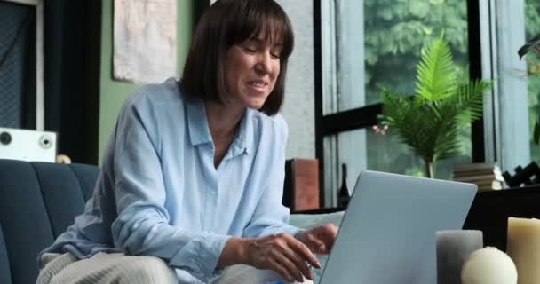 radosna i pozytywna biała kobieta rozjaśnia salon podczas rozmowy wideo za pomocą laptopa, siedząc na kanapie. Jej zaraźliwy optymizm promieniuje przez ekran.. - Materiał filmowy, wideo