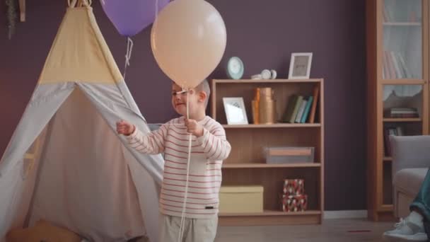 Medium shot of happy child with down syndrome throwing colorful party balloons in air while playing at home - Footage, Video