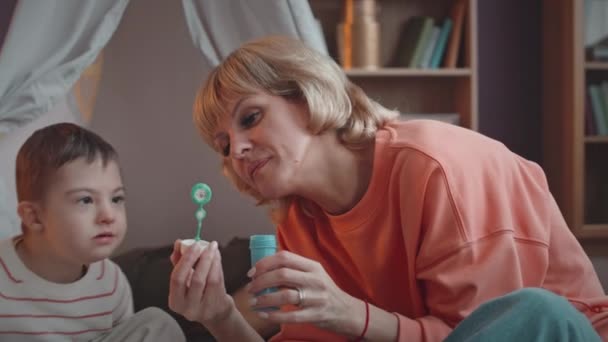 Lento de lindo niño caucásico de 6 años con síndrome de Down soplando burbujas de jabón con su madre en casa - Metraje, vídeo