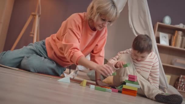 6-річний маленький хлопчик з синдромом Дауна грає з різнокольоровими будівельними блоками різної форми разом зі своєю коханою мамою, сидячи на підлозі в затишній вітальні - Кадри, відео