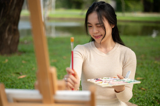 Una giovane artista asiatica bella e creativa sta misurando la sua immagine con un pennello, godendo la pittura su una tela mentre si rilassa in un parco verde. - Foto, immagini