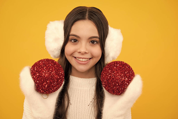 Merry christmas and happy New year. Portrait of happy teenager child wears sweater and knitted gloves on yellow isolated background. Kid in winter clothes. Teen girl with decorative christmas ball - Fotó, kép