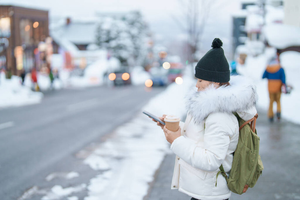携帯電話を使用している女性や、ニセコ旅行中の冬に雪の降る熱いコーヒーや紅茶を飲む女性。北海道の観光スポットとして人気のランドマーク。旅行と休暇の概念 - 写真・画像