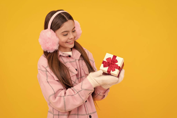 lachend kind in oorkappen en wanten houden geschenkdoos op gele achtergrond, kopen. - Foto, afbeelding