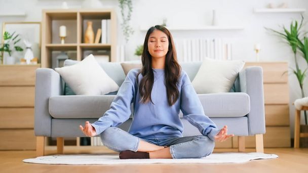 Счастливая молодая азиатская женщина практикует йогу и медиацию дома, сидя на полу в гостиной в позе лотоса и расслабляясь с закрытыми глазами. Заботливая концепция медитации и благополучия - Фото, изображение
