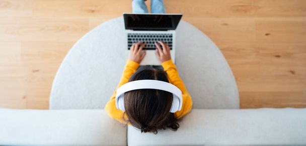 Top view of young Asian woman sitting on the floor and working on computer laptop, wearing wireless headset, listening to music - Photo, Image