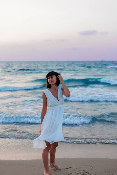Счастливая женщина в белом платье наслаждается отдыхом на пляже, гуляя вдоль песчаного берега на закате. счастливая девушка на пляже. - Фото, изображение