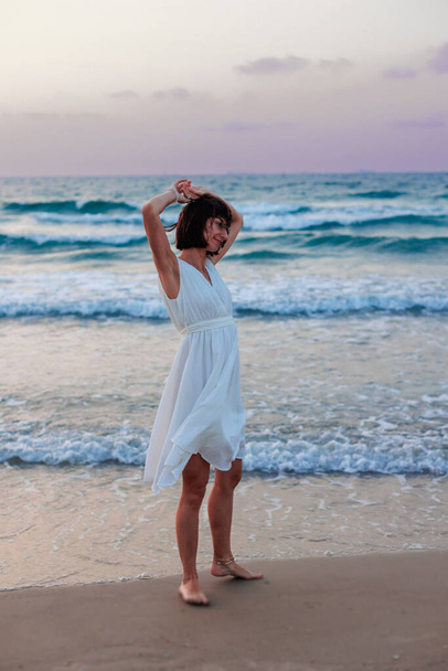 Szczęśliwa dziewczyna podróżnik w białej sukience letniej korzystających z tropikalnej plaży raj z turkusowym morzu. Szczęśliwa młoda kobieta w pięknej białej sukience spaceruje wzdłuż plaży podczas zachodu słońca.  - Zdjęcie, obraz
